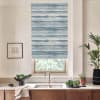 Blue-and-white watercolor-striped roller blind shields a kitchen window, softening daylight; sink and chrome faucet sit below on a marble countertop, flanked by potted plants and a framed sketch.