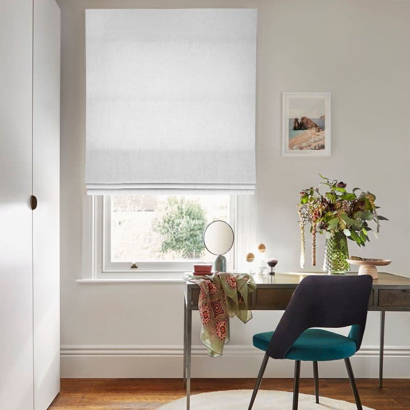 White linen Roman blind, fully lowered with subtle folds, filtering soft daylight through the window; compact home office with wooden desk, teal chair, a vase of flowers, and framed seaside print.