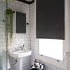 Black roller blind, lowered with visible beaded pull, blocking light through a window; small white subway-tiled bathroom with pedestal sink, mirror, hanging plant, and toiletries on folded towel.