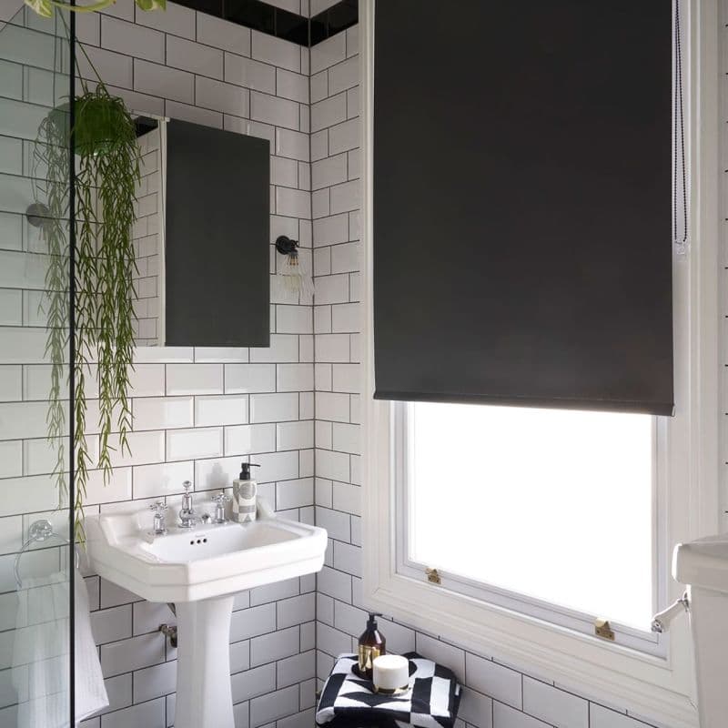 Black roller blind, lowered with visible beaded pull, blocking light through a window; small white subway-tiled bathroom with pedestal sink, mirror, hanging plant, and toiletries on folded towel.