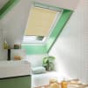 Beige pleated cellular blind lowered over a skylight, filtering daylight through a narrow lower gap; in a bright attic bathroom with green window trim, white herringbone tiles, bathtub, sink, plant.