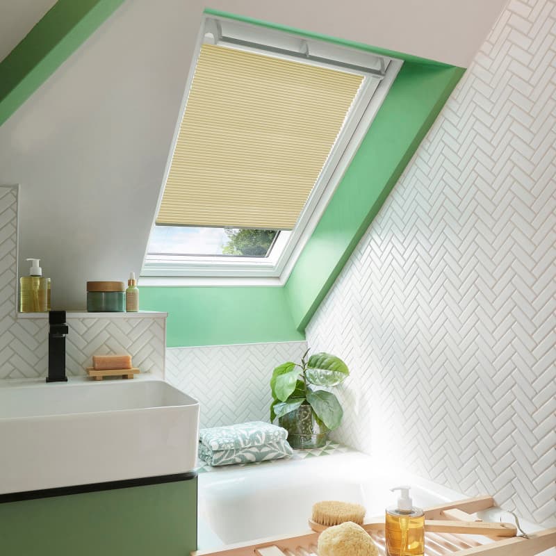 Beige pleated cellular blind lowered over a skylight, filtering daylight through a narrow lower gap; in a bright attic bathroom with green window trim, white herringbone tiles, bathtub, sink, plant.