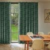 Green patterned, floor-length pleated curtains, mostly closed with a narrow central parting, filtering daylight and revealing a grassy backyard tree; mid-century dining room table, beige chairs and teapot nearby.