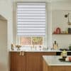 A dual-layer horizontal striped roller blind, lowered three-quarters, filtering daylight. Context: a kitchen with marble countertops, wooden cabinets, brass faucet, and a small brick-wall garden view.