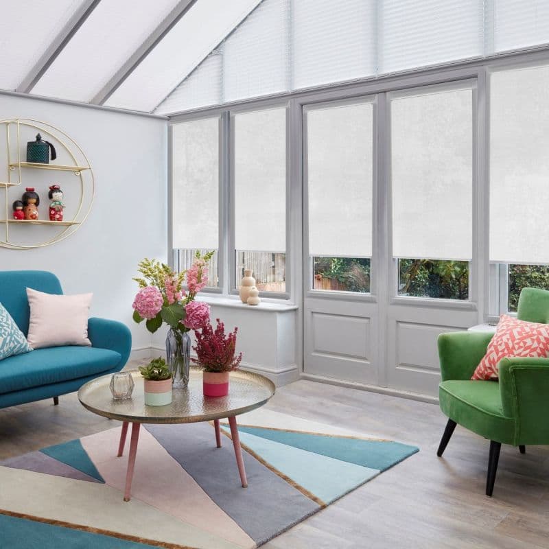 White roller blinds across tall conservatory windows and triangular roof panes, lowered to mid-height and diffusing light; bright sunroom with teal sofa, green armchair, round coffee table, pastel rug.