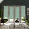 Glass French doors with teal roller blinds cover a living-room opening; blinds lowered over panes, framed by dark walls, green armchairs, glass coffee table on chevron rug, garden visible.