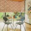 Bamboo-patterned fabric Roman blinds, lowered with soft folds and slightly raised bottom, filtering daylight. In sunlit dining nook with round tulip table, four wire chairs, vases, and garden view.
