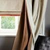 Roman blind with red fringe lowered, flanked by brown blackout and shimmery cream drapes pulled to the side; filtering soft daylight through window onto grey wall and woven leather chair.