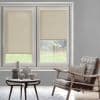 Beige roller blinds, both lowered (left fully, right partially), filtering daylight through two tall windows; muted living room with round wooden table and ceramics, rattan-leather armchair and grassy yard beyond.