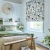 Patterned Roman blind over window, lowered and filtering daylight; geometric teal, blue and gray shapes on pale background. Bedroom setting with pale green bedding, white paneled wall, wooden bench, plants.