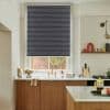 Dark pleated roller blind, fully lowered across a tall window, blocking most light, above a kitchen sink with brass faucet, white countertop, wooden cabinets and a brick courtyard visible below.