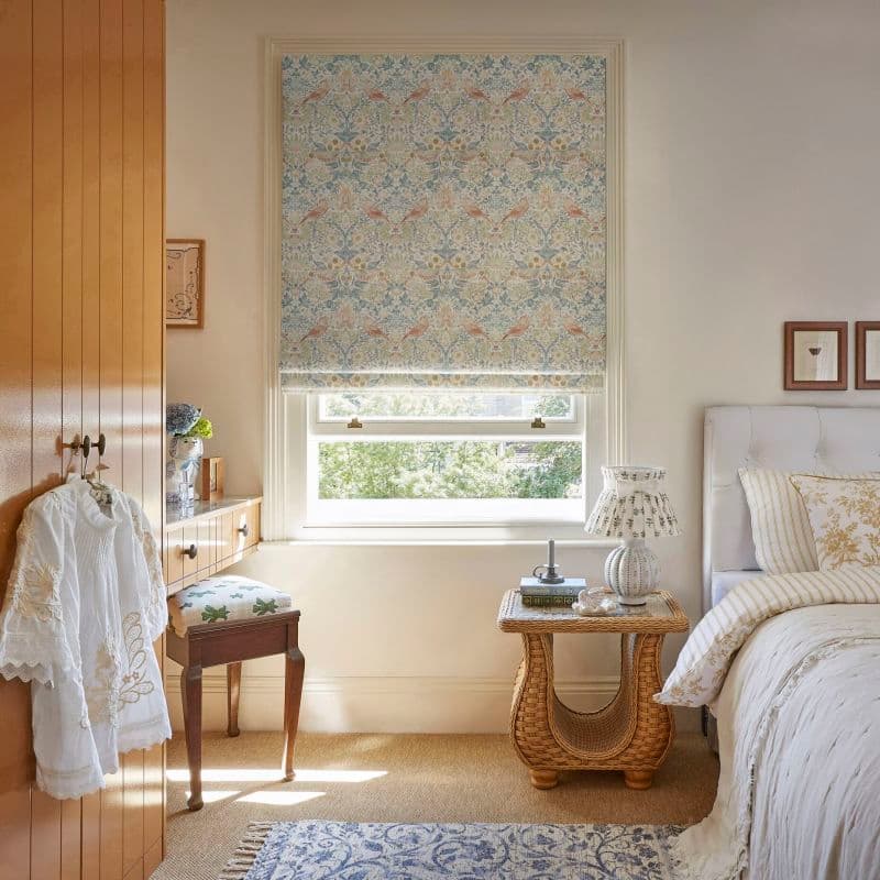 Patterned Roman blind with pastel floral-and-bird motif, lowered over the window, filtering soft daylight and revealing lower sash; cozy, neutral-toned bedroom with wicker bedside table, upholstered bed, leafy outside view.