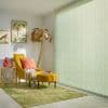 Pale green vertical blinds covering a full-height window, textured and drawn closed, softly diffusing sunlight. Cozy living-room corner with a mustard chaise lounge, coral cushion, gold palm lamp and bird prints.