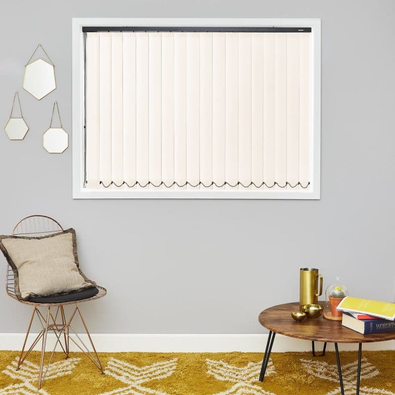 Vertical cream blinds covering a window, fully closed with decorative bead chain along the bottom, filtering soft daylight; mounted on a grey living-room wall above mustard rug, chair and table.