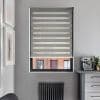 Zebra roller blind with alternating opaque gray and sheer horizontal bands, mostly lowered and filtering light over a window in a gray kitchen nook with radiator and espresso machine.