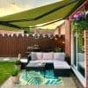 Green retractable awning with black metal arms, fully extended, shading a small backyard patio beside a brick house; wicker L-shaped sofa on tropical rug, hanging flowers, bunting at sunset.