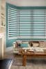 Dual-layer horizontal zebra blinds on a bay window, teal-and-sheer bands, lowered to filter light; set against teal walls above a beige sofa with cushions and a rustic wooden coffee table.