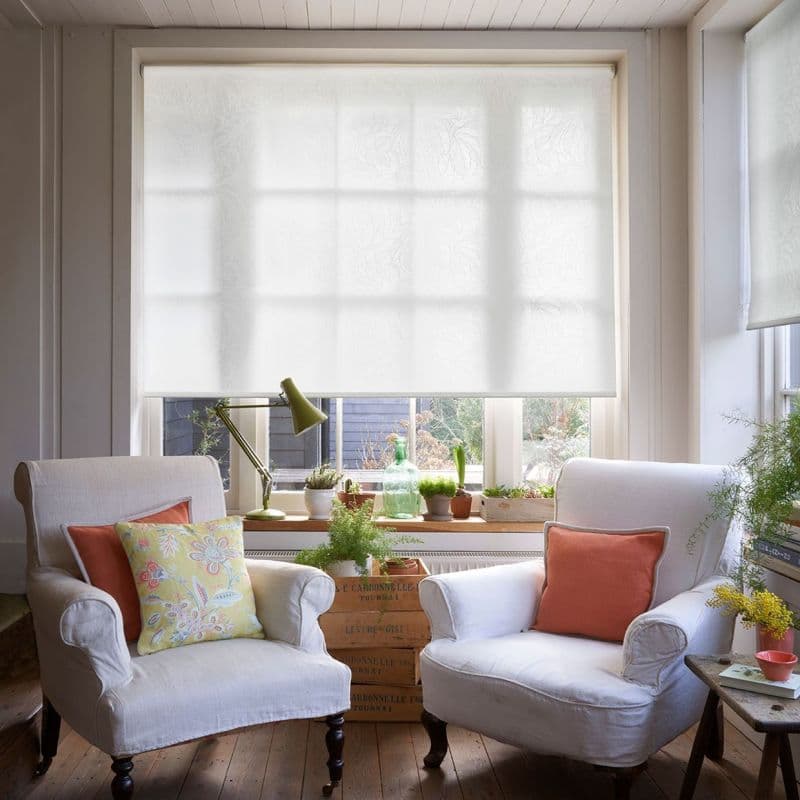 White translucent roller blind with subtle leaf pattern, lowered across a multi‑pane window and gently diffusing daylight; framed by a bright, cozy sitting room with two armchairs, plants, and wooden floor.