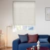 A cream-colored cellular blind covers the upper window, fully lowered with faint horizontal ribs, filtering daylight; below a small reveal shows a roof. Bright living room with blue sofa, cushions, lamp.