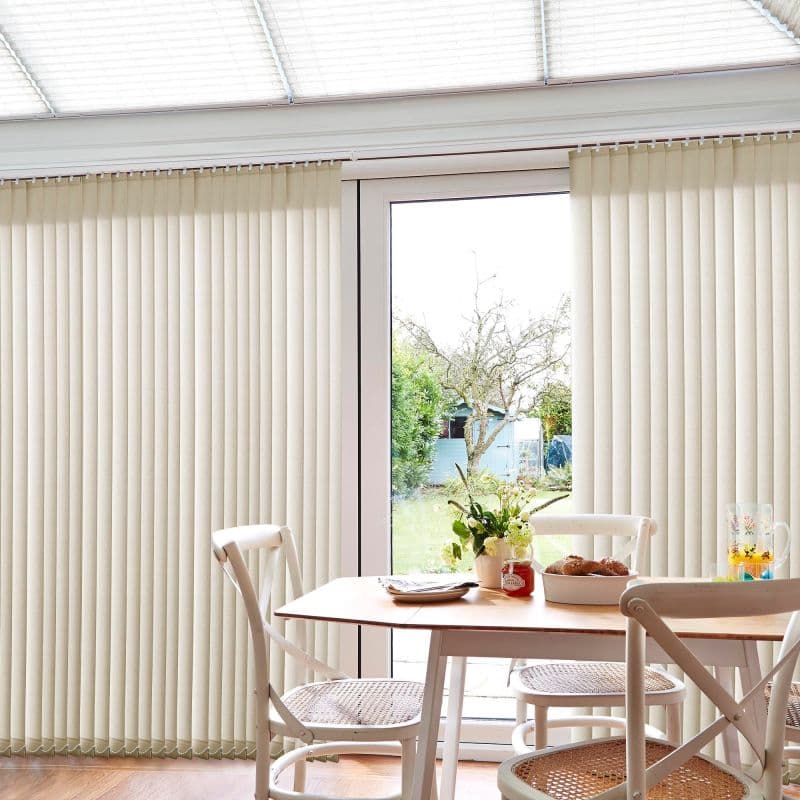 Cream vertical blinds across a sliding patio door, panels mostly closed with a central gap; they filter soft daylight and reveal a garden view, framing sunlit dining table and chairs.