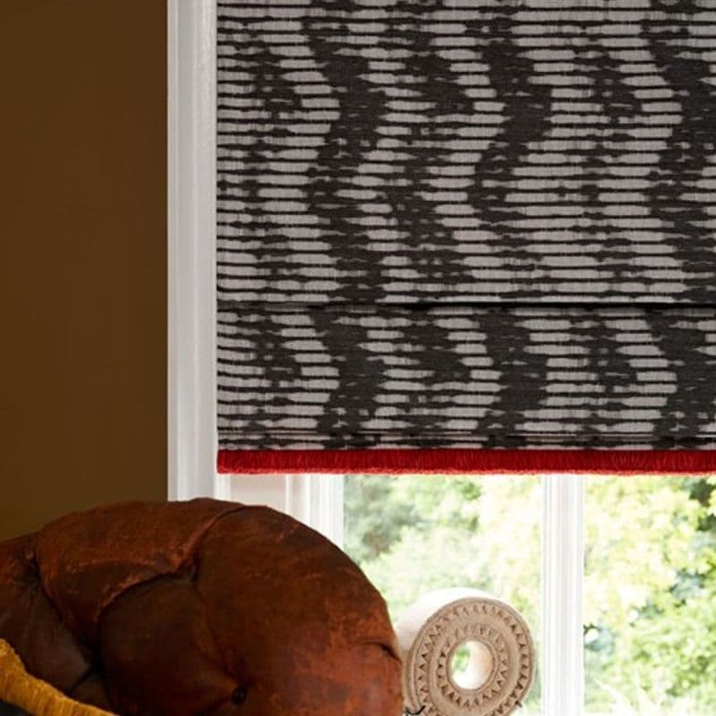 Roman shade with gray-and-white abstract horizontal pattern and red fringe trim, lowered over window and filtering daylight; living-room corner with brown leather chair arm, carved finial and leafy exterior view.