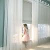 Sheer grommet-top curtains over a lowered roller blind, panels parted, filtering soft daylight as a girl in a dress and wings leans at the sill, bright room with garden view.