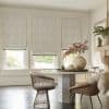Three patterned Roman shades covering three tall windows, partially lowered and filtering soft daylight, in bright neutral dining nook with round marble table, woven chairs, vase and garden view.
