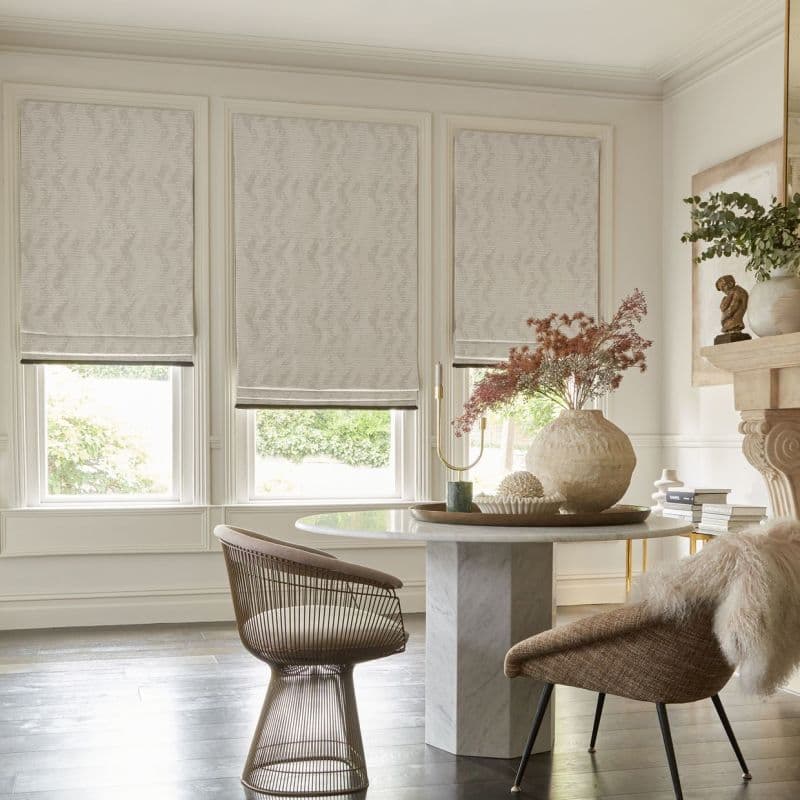 Three patterned Roman shades covering three tall windows, partially lowered and filtering soft daylight, in bright neutral dining nook with round marble table, woven chairs, vase and garden view.