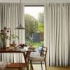 Full-length beige pleated curtains, hung floor-to-ceiling and parted at the center, framing sliding glass door; sunlight filters through onto wooden dining table set for breakfast with pendant lamp and garden view.