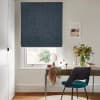 Dark-blue textured Roman blind lowered above the sill, filtering daylight over a window; small home desk scene with teal chair, mirror, vase of flowers and framed seaside print.