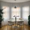 Sheer white floor-to-ceiling curtains on a curved bay of three windows, gently diffusing daylight; round marble-topped dining table with chairs and vase on a warm wooden floor.