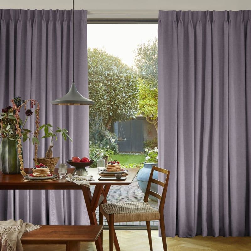 Floor-length pleated lavender-gray curtains on a track, drawn open at center to frame glass doors; admitting daylight into a dining area with table, pancakes, pendant lamp and sunlit garden.