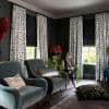 White patterned floor-length curtains flank three windows, paired with partially lowered black Roman shades that filter daylight; a dark-painted living room with velvet armchairs, plants and eclectic decorative objects.