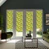 Four tall window panels fitted with yellow-and-green leaf-patterned roller blinds, filtering sunlight into a living room with green armchairs, glass coffee table, and a chevron rug.