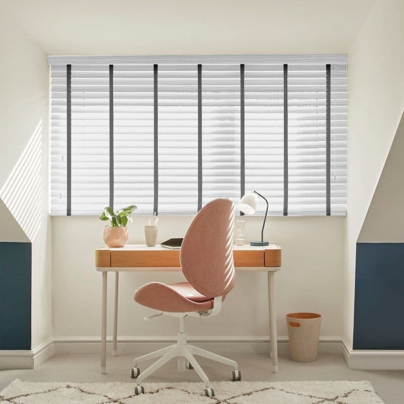 White horizontal blinds with dark vertical tapes covering a large dormer window, partially closed and filtering soft daylight, above a compact home office desk with pink swivel chair and lamp.