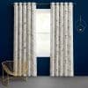 Grommet-top white floral curtains, pleated and hanging on a metal rod, frame a window and filter daylight; against a deep navy wall with gold wire chair on gray carpet.