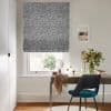 Gray-and-white geometric Roman blind lowered over the window, filtering daylight; above a wooden desk in a bright home office with a teal chair, vase of flowers, and framed seaside print.
