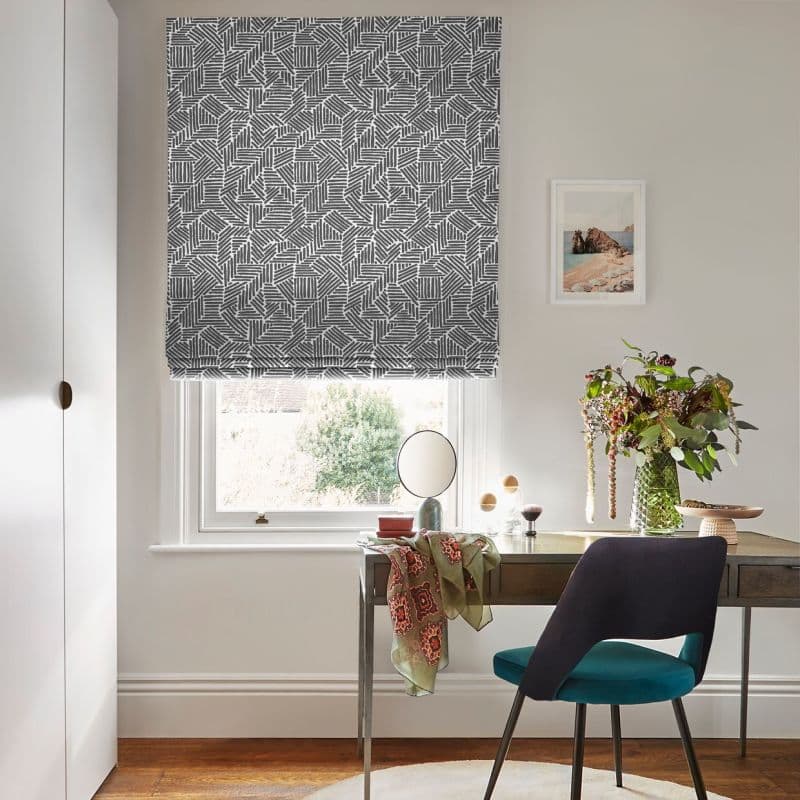 Gray-and-white geometric Roman blind lowered over the window, filtering daylight; above a wooden desk in a bright home office with a teal chair, vase of flowers, and framed seaside print.