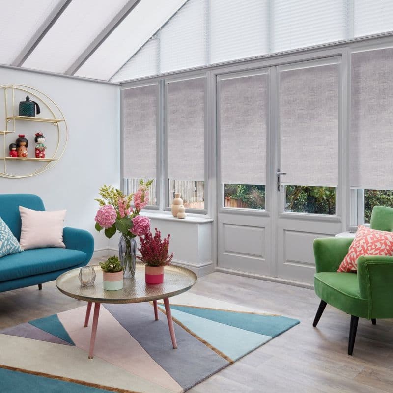 Light gray roller blinds covering French doors and lower window panes, partly lowered and filtering daylight; modern sunroom/living area with pastel furniture, round coffee table, floral arrangements and garden view.