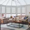 White pleated blinds on bay windows and conservatory roof, drawn to diffuse light; filtering soft daylight into a sunroom-style living area with neutral sofa, pink chunky throw, and wooden coffee table.