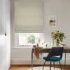 Roman fabric blind, light beige, fully lowered with soft horizontal folds, filtering daylight into a study; wooden desk, teal chair, mirror and vase of flowers beside a small framed seascape.