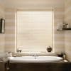 Horizontal beige Venetian blinds, fully lowered, filtering soft daylight; positioned over a window above a white oval bathtub in a softly lit bathroom with striped walls and toiletries.