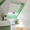 White pleated skylight blind, lowered halfway across a tilted window, filtering soft daylight; set in a green‑trimmed attic bathroom with white herringbone tiles, sink, tub, and a potted plant.