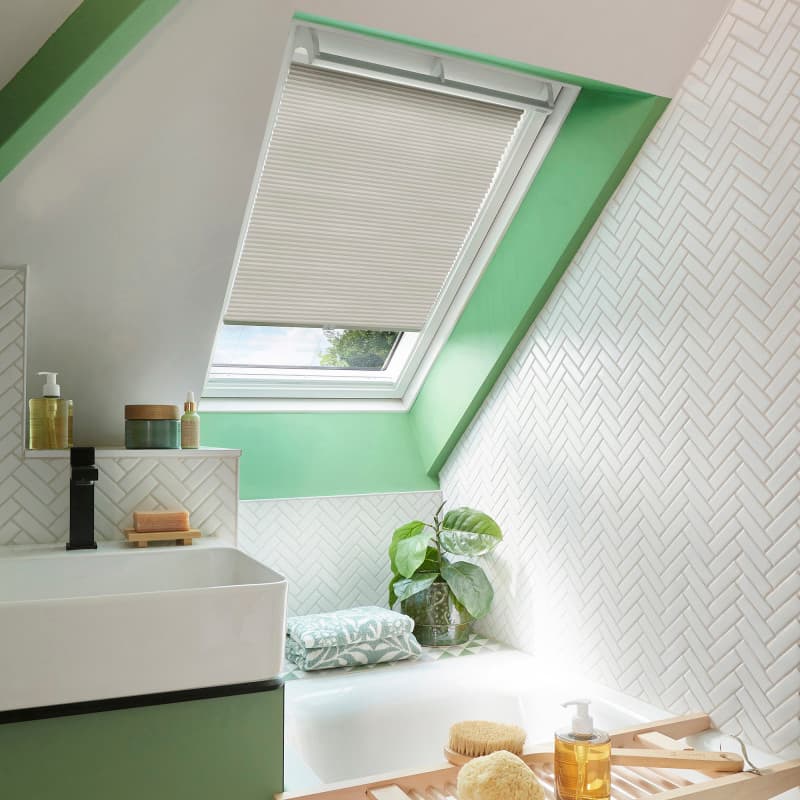 White pleated skylight blind, lowered halfway across a tilted window, filtering soft daylight; set in a green‑trimmed attic bathroom with white herringbone tiles, sink, tub, and a potted plant.