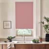Rose-colored roller blind hangs nearly fully lowered over a kitchen window, filtering light; below stainless faucet and double sink sit on stone countertop with wooden cabinets, plants and bowls.