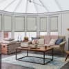 Pale green pleated cellular blinds cover tall conservatory windows, lowered mid-height and softly filtering daylight for privacy; framed by a beige sofa, wooden coffee table, and blue patterned rug.