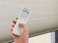 Cellular honeycomb blind lowered, filtering daylight; hand holds a white remote with thumb on its buttons, aiming at the window; interior with soft light and a tiled patio visible outside. Text: 8 somfy
