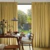 Floor-length pleated golden curtains, mostly drawn with a central opening, gently filtering daylight. They frame a dining table set for breakfast, pendant lamp overhead, and a lush backyard garden beyond.