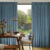 Blue pleated curtains, parted at center to admit daylight through sliding glass doors; dining table set for breakfast with wooden chairs, pendant light, and a sunlit garden visible beyond.