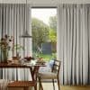 Pleated floor-to-ceiling light-gray patterned curtains, parted at center and partially drawn, filtering morning light onto a breakfast-set wooden table with chairs and pendant lamp, opening to a sunlit garden view.